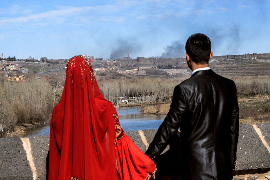 Ein Hochzeitspaar posiert für ein Foto. Im Hintergrund sind Rauchsäulen über der Stadt Diyarbakir zu sehen, in der regelmäßig türkische Truppen und kurdische Rebellen aufeinander treffen