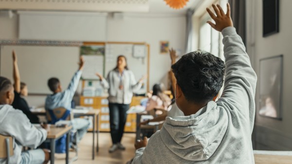 In einem Klassenraum sieht man aus Rückenansicht einen jungen Schüler, der sich meldet.