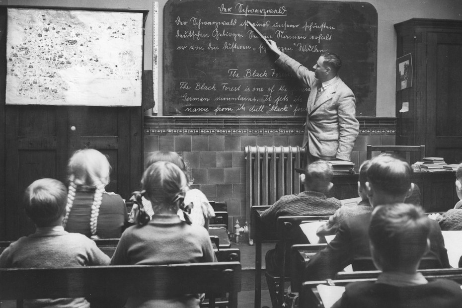 Ein Lehrer erteilt eine Unterrichtsstunde in Englisch und Deutsch. Die Kinder sitzen nebeneinander und haben so den Vorteil, die Sprache des anderen zu lernen (1930).