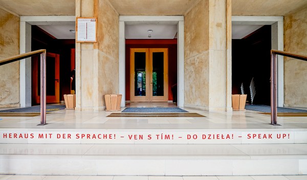 Eine weiße Treppe, die zum Eingang einer braunen Tür führt. Auf der weißen Treppenstufe steht in roter Schrift: „Heraus mit der Sprache“ in verschiedenen Sprachen.
