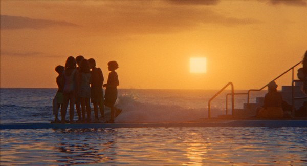 Eine Gruppe junger Menschen steht auf einem Steg auf dem Wasser. Dahinter sieht man den Sonnenuntergang. Das Bild ist leicht unscharf.